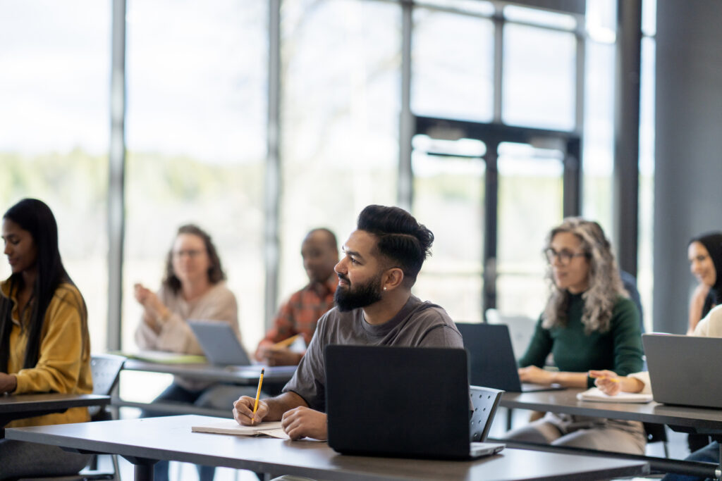A large group of mature adults are seen sitting at desk with wither laptops and books open as they study during a continuing education class. They are each dressed casually and are listening attentively.