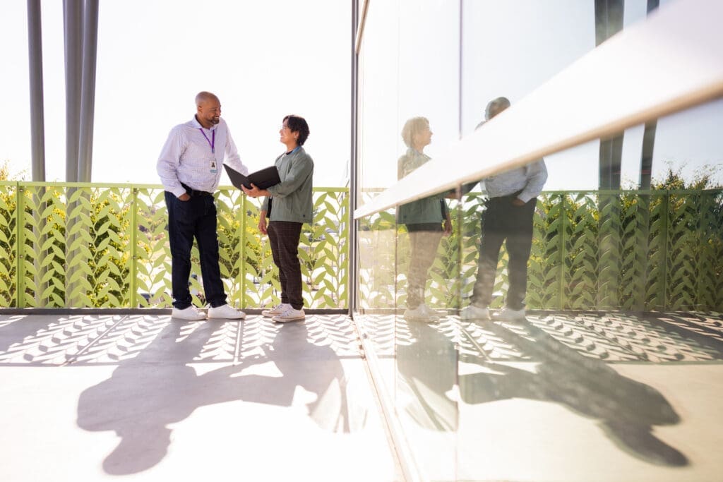 Two individuals conversing on a sunny balcony with a leafy green railing, one holding a black folder. Their reflections can be seen on a glass wall.