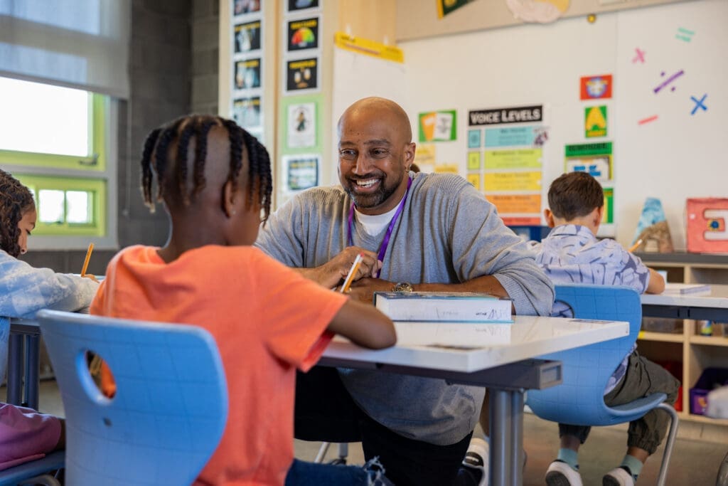 A teacher interacts with a young student in a bright classroom, while two other students work at their desks.