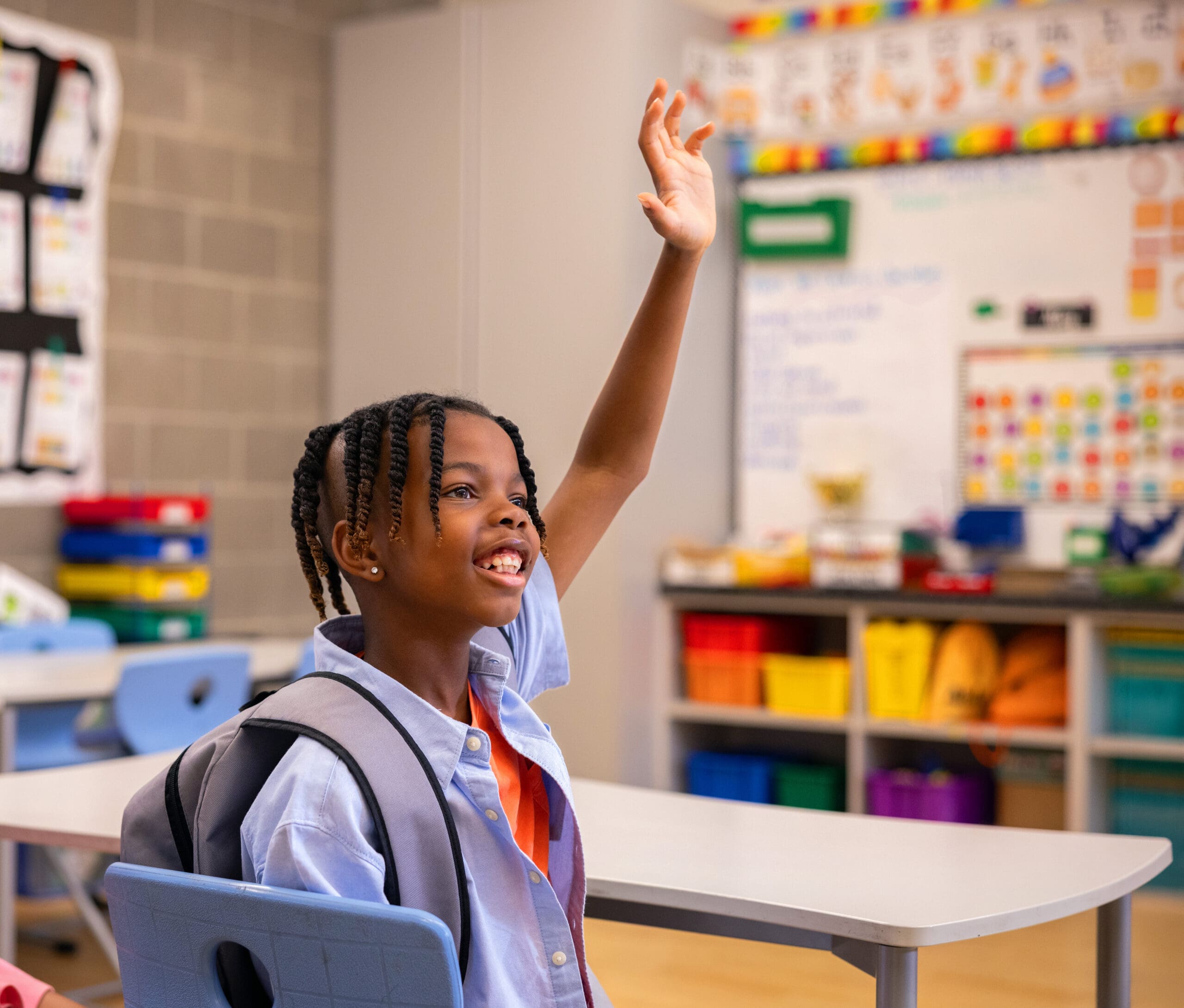 A smiling girl with braided hair raises her hand in a classroom, wearing a blue shirt and gray backpack.