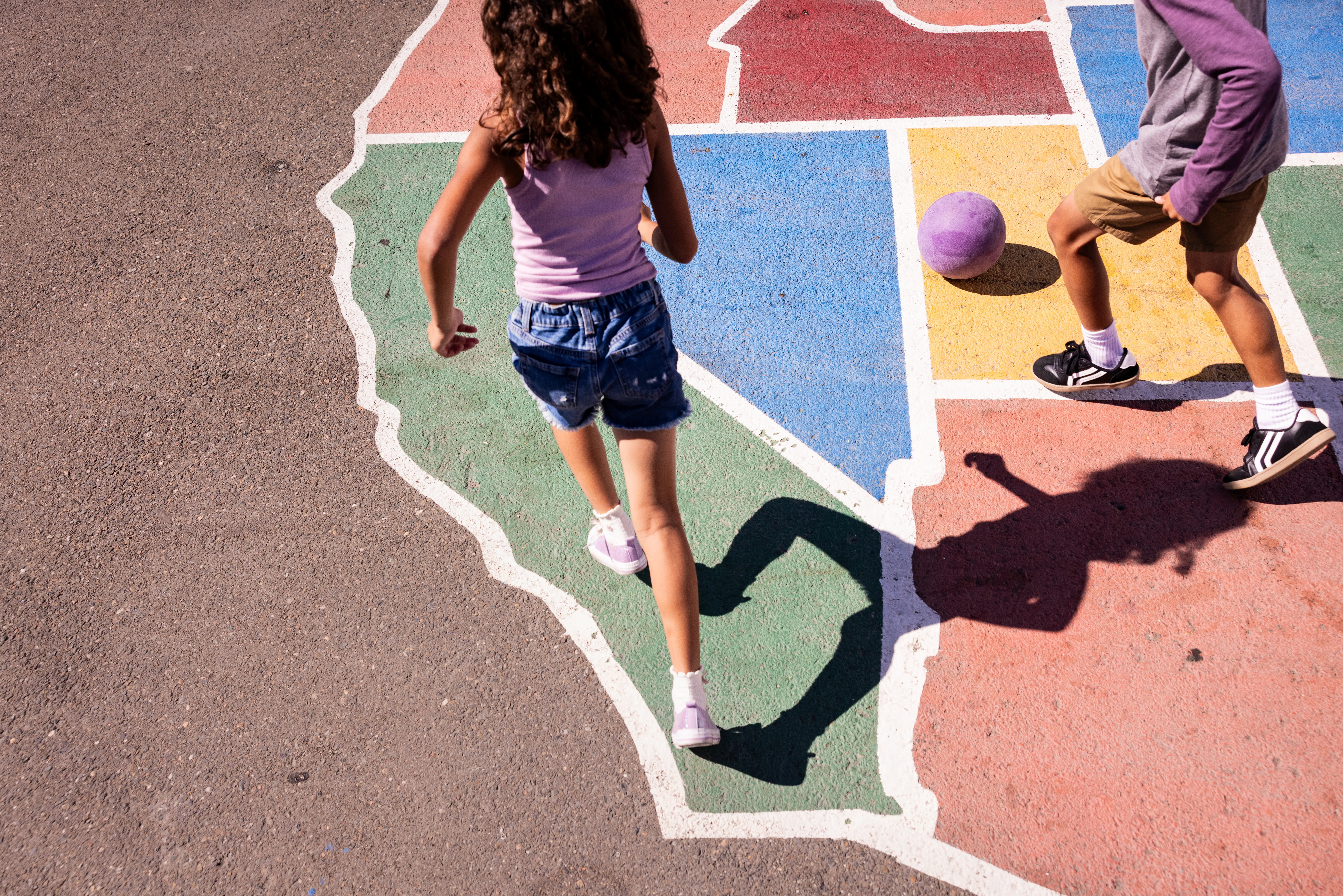 Children play on a colorful asphalt map of the United States, with one girl running and a boy kicking a purple ball.